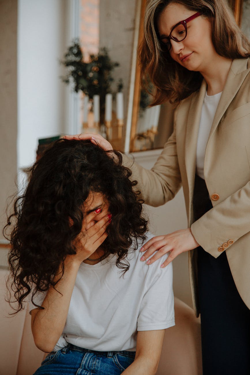 A woman in a beige blazer places her hand gently on another person's head, leaning in close with full attention.
