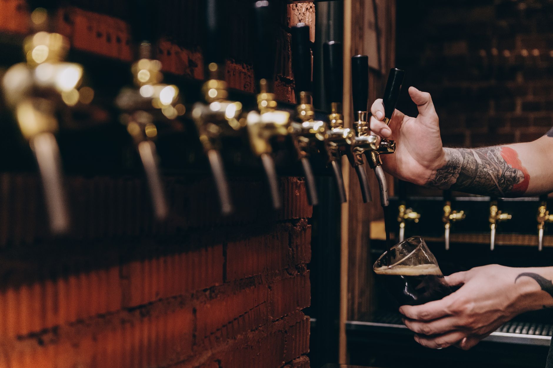 A bartender pours a draft beer at a dimly lit microbrewery, the tap handles glowing gold in the low light.
